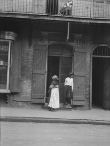 View from across street of a man and woman standing by a doorway and a woman seated...c1920-1926. Creator: Arnold Genthe.