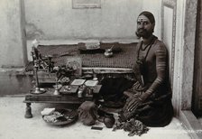 An Indian man wearing elaborate jewellery and make-up, squatting in front of a small table..., c1900 Creator: Unknown.