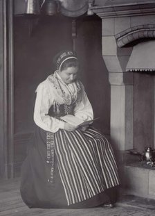 A woman sits in front of a fireplace and reads dressed in a Dalarna costume, 1886-1920. Creator: Helene Edlund.