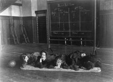 Five girls lying on mat in gymnasium in front of basketball scoreboard..., Washington, D.C., (1899?) Creator: Frances Benjamin Johnston.
