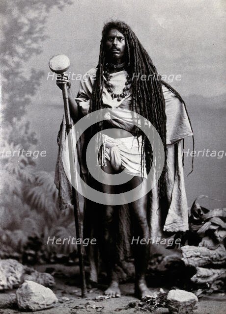 An Indian man with long hair, carrying a staff, in a studio setting, 1900. Creator: Unknown.