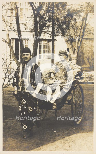 A man in ornate drag sits in a dog-cart and is pulled along the road by...(between 1910 and 1919?). Creator: Unknown.