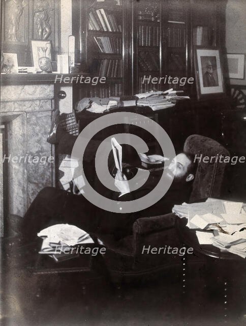Arthur Schuster, lying on an armchair by a fireplace, with a photograph of Lord Kelvin..., c1901. Creator: Unknown.