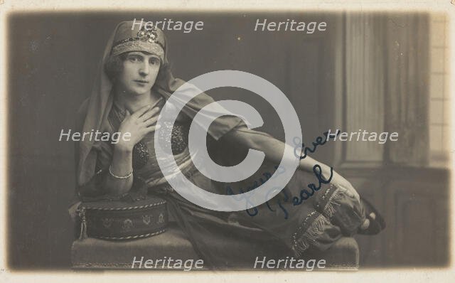 A man in drag, poses wearing delicate attire; curled up on a piece of furniture, [1910-1919?]. Creator: Unknown.