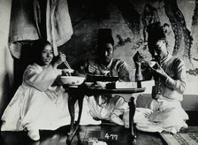 A group of people eating a meal, seated behind a small table, in front of a painted screen, c1900. Creator: Unknown.