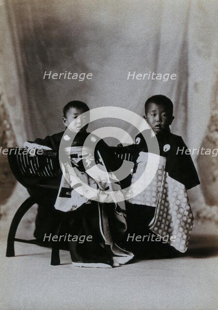Two boys in a photographic studio, one seated on a chair, in front of a curtained backdrop, c1900. Creator: Unknown.