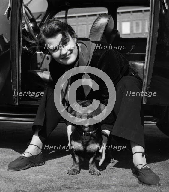 Photographer Kerstin Bernhard sits in the doorway of her car and holds a dachshund, 1955-1960. Creator: Unknown.
