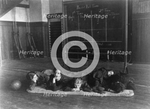 Five girls lying on mat in gymnasium in front of basketball scoreboard..., Washington, D.C., (1899?) Creator: Frances Benjamin Johnston.