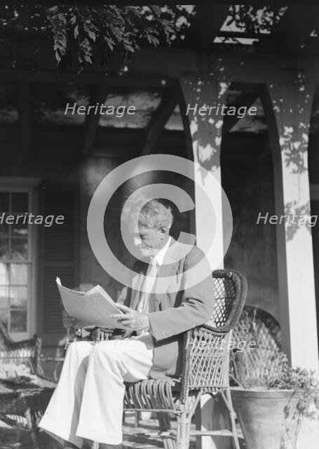 Arnold Genthe seated outdoors in a wicker chair reading, between 1911 and 1942. Creator: Arnold Genthe.