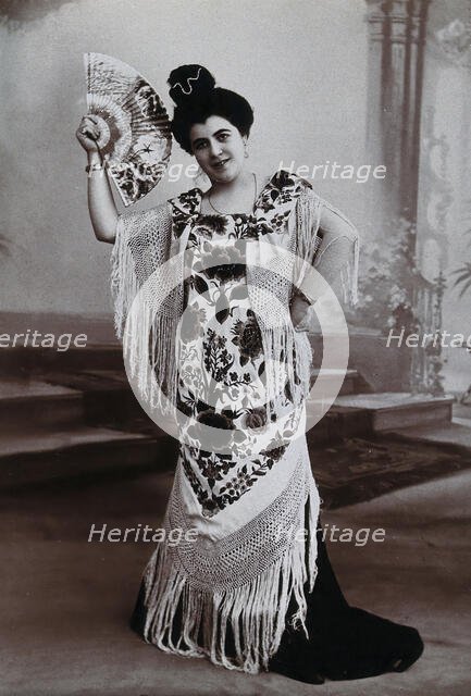 A standing woman, holding aloft a fan, wearing a fringed shawl, in front of a painted..., c1900. Creator: Unknown.