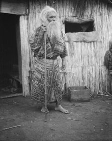Ainu man holding a staff standing outside a hut, 1908. Creator: Arnold Genthe.