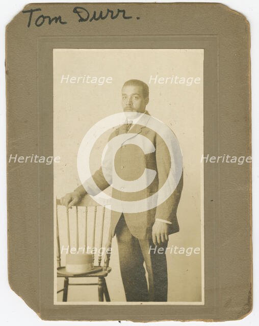 Photograph of Tom Durr standing next to a chair with a hat on it, ca. 1910. Creator: Unknown.