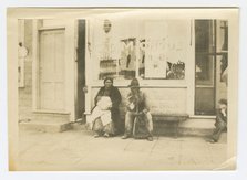 Photograph of a man and woman sitting outside of a storefront, early 20th century. Creator: Unknown.