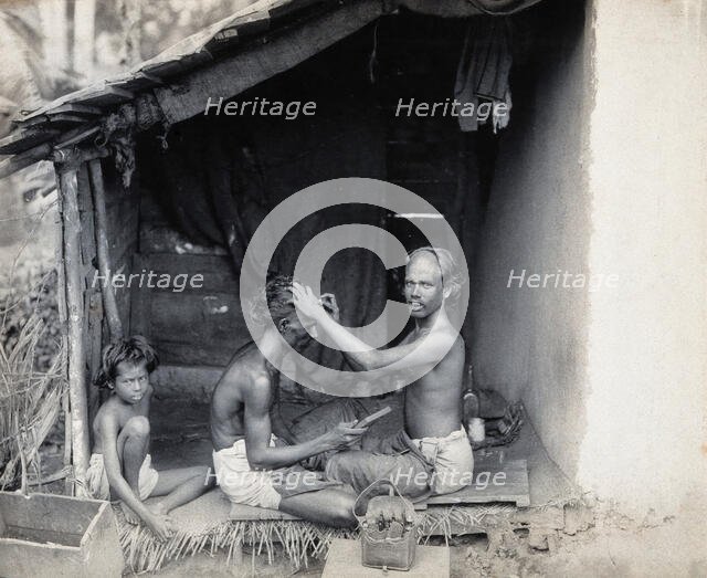 Two men sitting cross-legged; one is a barber shaving the front of the other's head, c1900. Creator: Unknown.