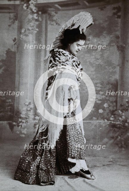 A standing woman, posed with an upraised fan and pointed toe, wearing a fringed shawl, in..., c1900. Creator: Unknown.
