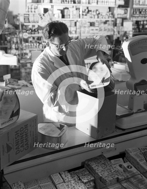 Shopkeeper unpacking canned gammon joints, Mexborough, South Yorkshire, 1963.  Artist: Michael Walters