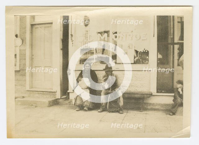 Photograph of a man and woman sitting outside of a storefront, early 20th century. Creator: Unknown.