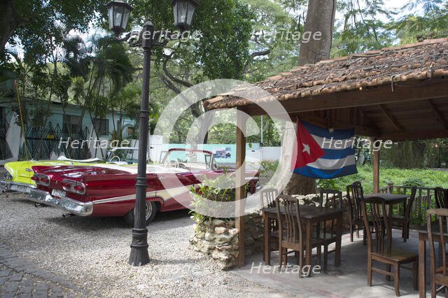 Old American cars and the Cuban flag at the Bosque, Havana, Cuba, 2024. Creator: Ethel Davies.