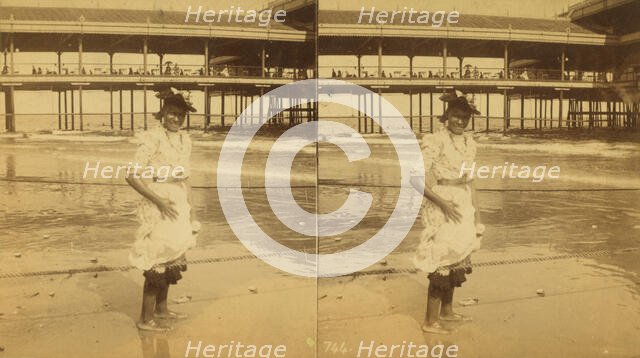 Young woman wading at the beach, in front of a covered pier, (1868-1900?). Creator: Unknown.