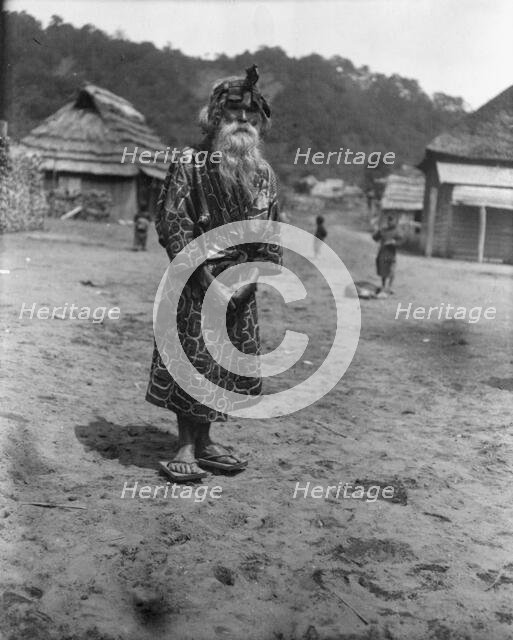 Ainu chief wearing a headdress standing in the village lane, 1908. Creator: Arnold Genthe.