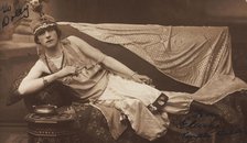 A man in drag poses wearing delicate attire; curled up on a piece of furniture, [1910-1919?]. Creator: Unknown.