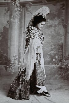 A standing woman, posed with an upraised fan and pointed toe, wearing a fringed shawl, in..., c1900. Creator: Unknown.