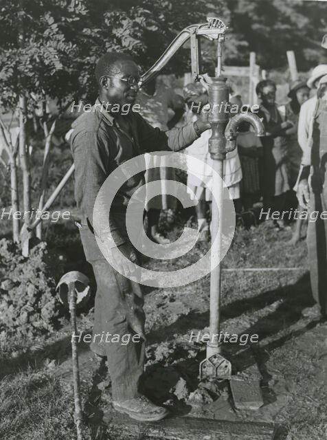 An African American man fixing a well's tube into the ground near Ridge, Maryland, July 1941. Creators: Farm Security Administration, John Collier.