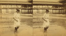 Young woman wading at the beach, in front of a covered pier, (1868-1900?). Creator: Unknown.
