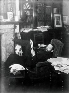 Arthur Schuster, lying on an armchair by a fireplace, with a photograph of Lord Kelvin..., c1901. Creator: Unknown.