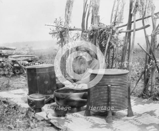 Ainu man seated outdoors on a mat covered with clay containers, 1908. Creator: Arnold Genthe.
