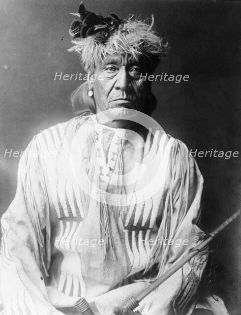 Atsina Indian, Red Whip, half-length portrait, seated, facing front, wearing feather..., c1908. Creator: Edward Sheriff Curtis.