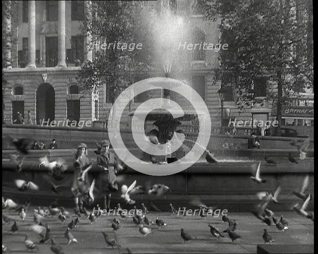 A Boy and a Girl Feeding Pigeons by One of the Fountains in Trafalgar Square with..., 1939. Creator: British Pathe Ltd.