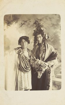 Two men in drag, one wearing a detailed feathered head garment, pose for a portrait, [1900-1909?]. Creator: Frank Wood.