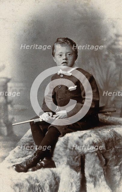 A boy with Down's syndrome, holding a rifle and sitting on a fur carpet, c1900. Creator: RW Morris.