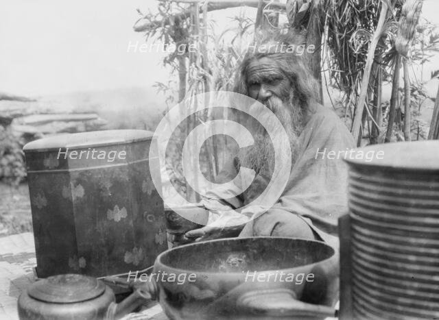Ainu man seated outdoors on a mat covered with clay containers, 1908. Creator: Arnold Genthe.
