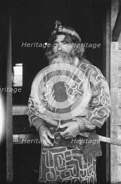Ainu chief wearing a headdress and holding a sword standing in a doorway, 1908. Creator: Arnold Genthe.