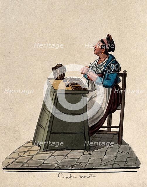 A women is sitting knitting at a table in the street with money on it in trays. Creator: Unknown.