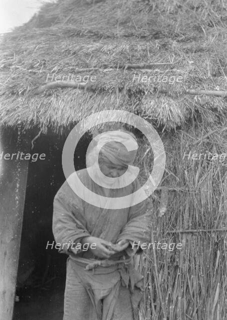 Ainu woman standing outside the entrance of a hut, 1908. Creator: Arnold Genthe.