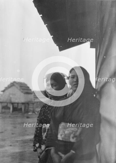 Two Ainu women standing outside by the wall of a wooden hut, 1908. Creator: Arnold Genthe.