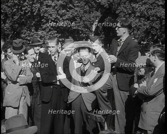 A Group of Male Civilians and One Soldier Listen to a Man With a Cigarette Standing on a..., 1938. Creator: British Pathe Ltd.