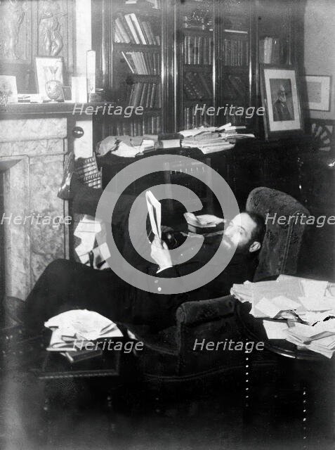 Arthur Schuster, lying on an armchair by a fireplace, with a photograph of Lord Kelvin..., c1901. Creator: Unknown.