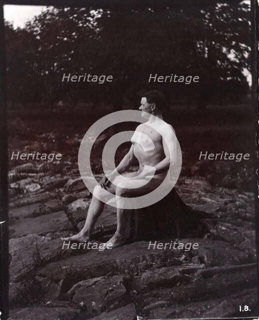 A man posing naked, seated on a rocky outcrop in a leafy landscape, c1900. Creator: Unknown.
