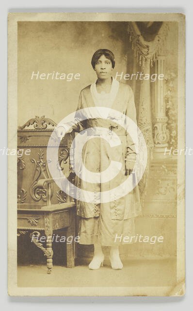 Photographic postcard of a woman standing next to a wooden chair, 1904-1918. Creator: Unknown.