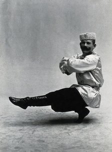 A man, in a satin tunic and fur hat, dancing a "Cossack" measure in a studio setting, 1899-1908. Creator: Unknown.