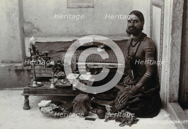 An Indian man wearing elaborate jewellery and make-up, squatting in front of a small table..., c1900 Creator: Unknown.