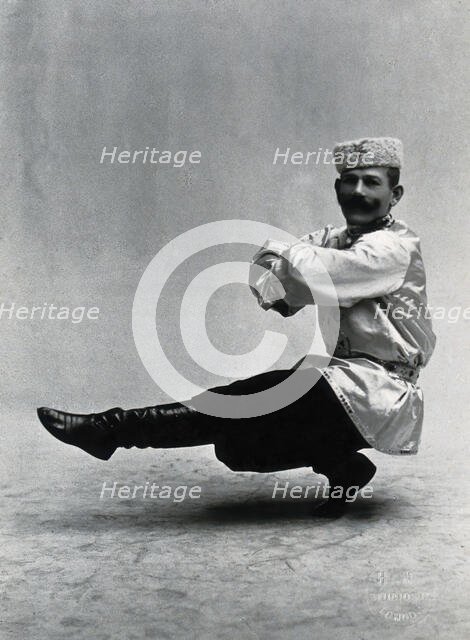 A man, in a satin tunic and fur hat, dancing a "Cossack" measure in a studio setting, 1899-1908. Creator: Unknown.