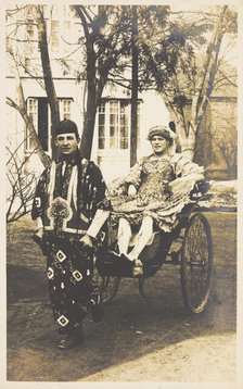 A man in ornate drag sits in a dog-cart and is pulled along the road by...(between 1910 and 1919?). Creator: Unknown.