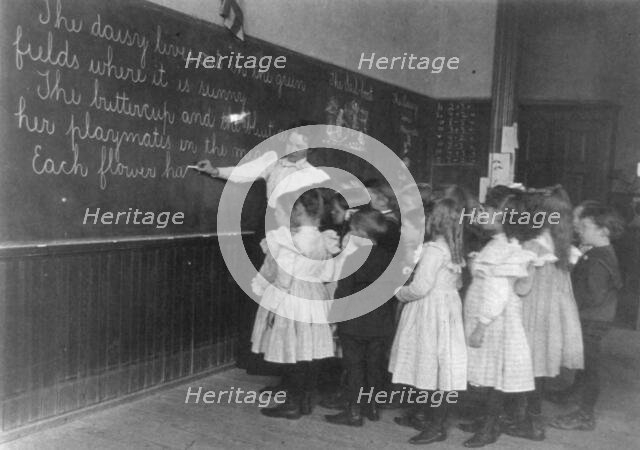 Elementary school children standing and watching teacher write at blackboard, Washington DC, (1899?) Creator: Frances Benjamin Johnston.