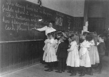 Elementary school children standing and watching teacher write at blackboard, Washington DC, (1899?) Creator: Frances Benjamin Johnston.