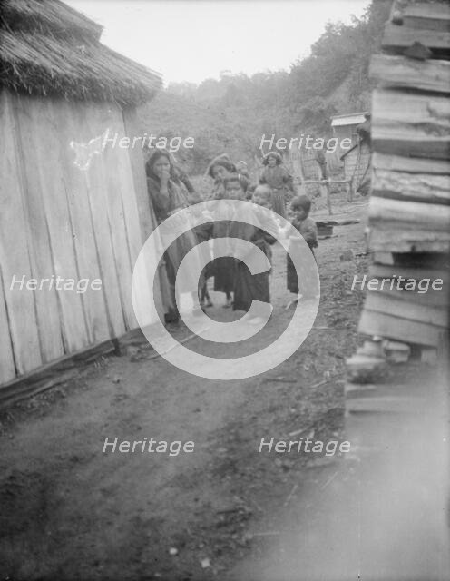 Group of Ainu children standing in a passageway between huts, 1908. Creator: Arnold Genthe.